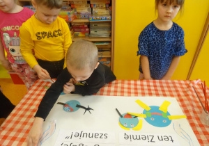 Trójka dzieci dekoruje plakat ekologiczny, naklejają elementy z kolorowego papieru.
